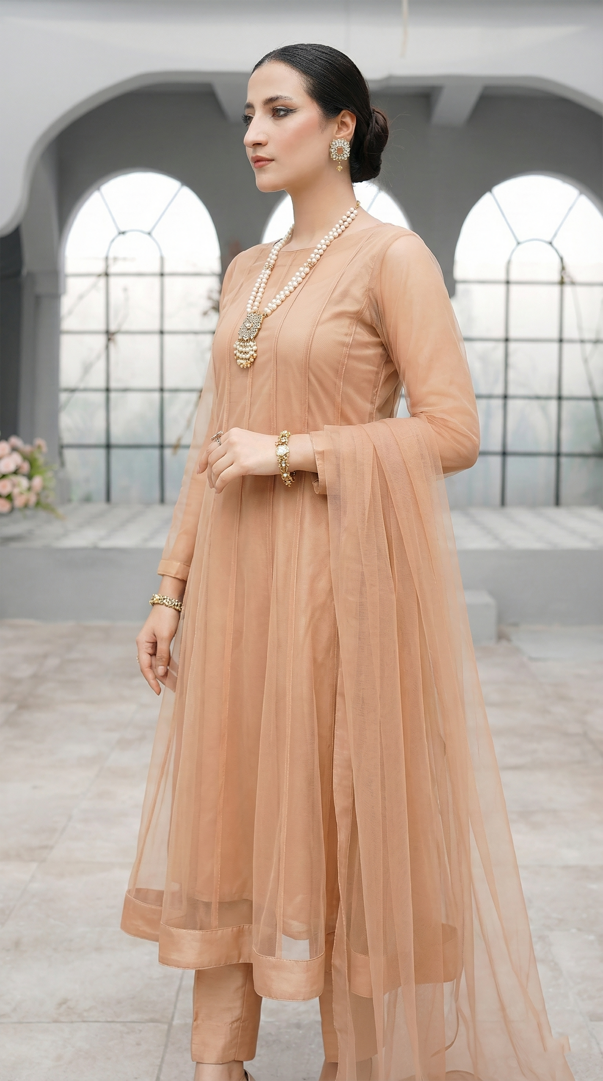 ROZAIB Champagne Peach luxury Bareeze net minimal kalidaar stitched suit. This premium ready to wear stitched dress features a 12-kali graceful flow fit, ultra-fine 0.3mm micro-mesh texture, and master-tailored vertical kali joints. A minimalist 3-piece stitched suit for women in a warm champagne-peach tone, paired with raw silk cigarette trousers and an airy Bareeze net dupatta. Designed for a traditional yet modern festive look with zero embroidery.